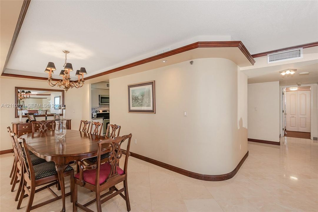 Curved architectural wall feature, polished stone-finish tile flooring, custom tiered wood crown molding, and ornate multi-arm chandelier