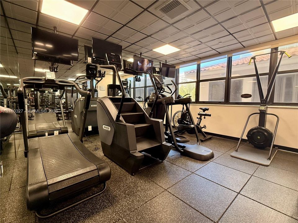 Fitness center featuring rubber flooring, mirrored walls, recessed grid ceiling lighting, and large windows with horizontal blinds