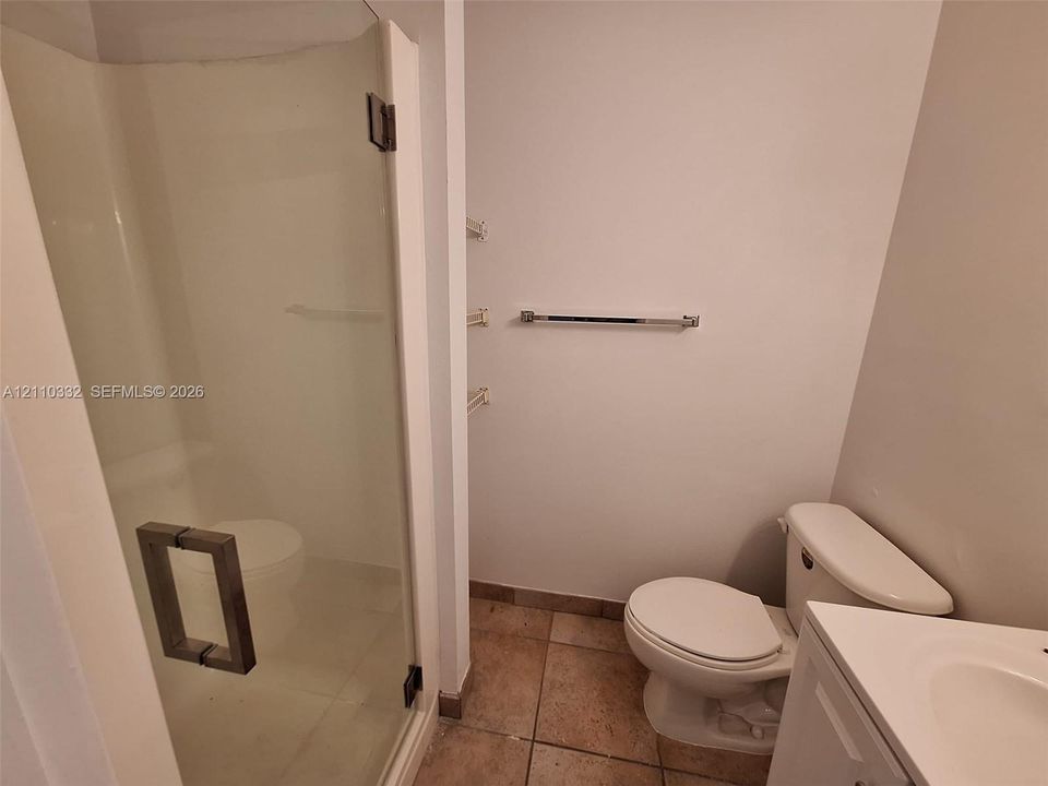 Walk-in shower with clear glass enclosure and square metallic pull handle, ceramic tile flooring, white vanity with integrated sink, and wall-mounted metal towel bar