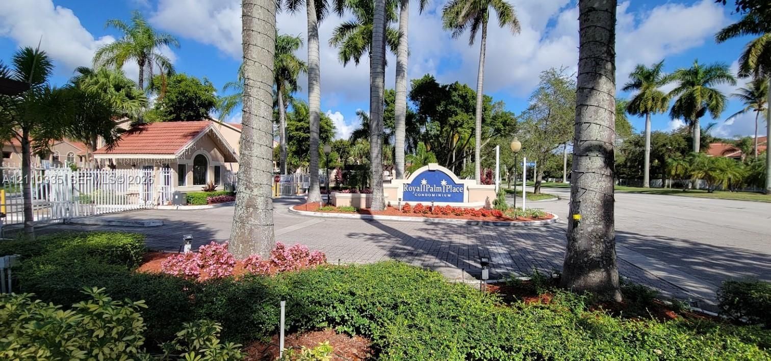 Gated community entrance featuring decorative masonry signage, paved access road, white metal perimeter fencing, and mature palm landscaping