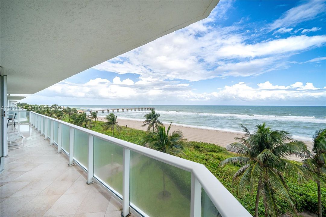 Covered balcony featuring tile flooring and a glass-paneled railing overlooking the ocean, sandy beach, and palm trees