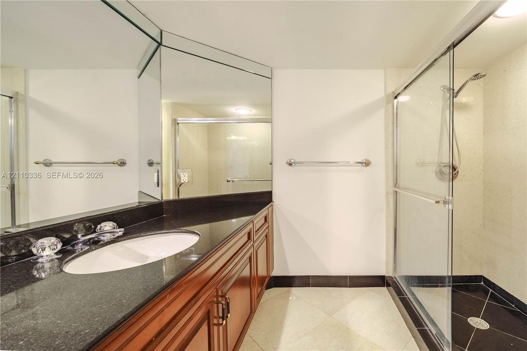 Vanity with dark countertop and white oval under-mount sink, wood-finish cabinetry, floor-to-ceiling mirror, and glass-enclosed shower with tiled flooring