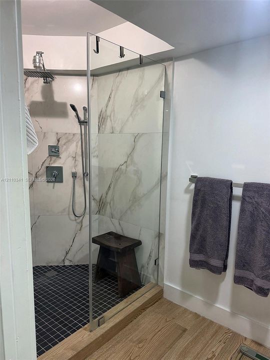Walk-in shower featuring large-format stone-pattern tile walls, a grid-patterned floor, a frameless glass door, a rainfall showerhead with a handheld attachment, and wood-finish flooring