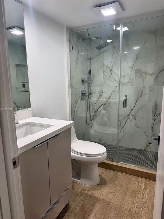 Bathroom featuring a vanity with a white countertop and rectangular sink, frameless glass-enclosed shower with large-format stone-patterned wall tile, rain-style shower head, and wood-finish flooring