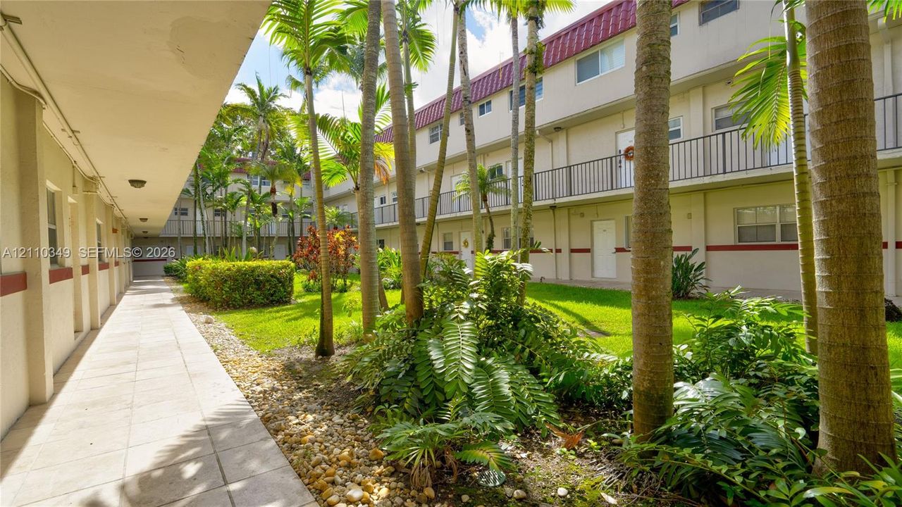 Covered exterior walkway featuring a tiled floor, manicured lawn, stone ground cover, mature palm trees, and multi-level railings