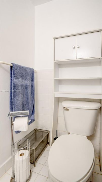 White tile wall surround, porcelain commode, and built-in white over-the-toilet storage unit featuring dual cabinet doors and open shelving