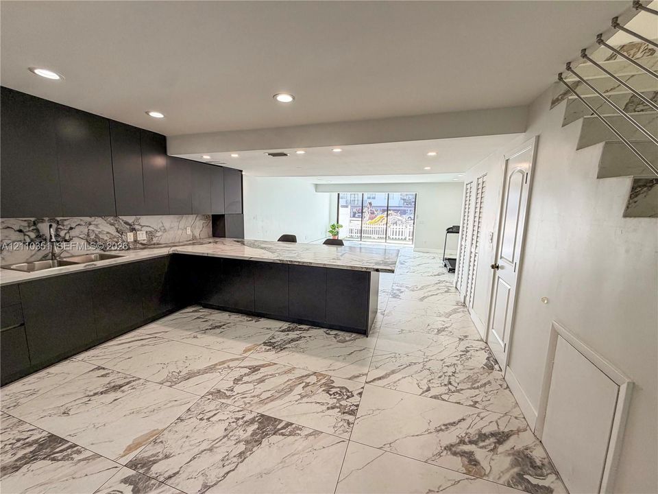Modern kitchen featuring flat-panel matte cabinetry, extensive stone-patterned surfacing, large-format tile flooring, integrated recessed lighting, and a floating staircase with metal railings