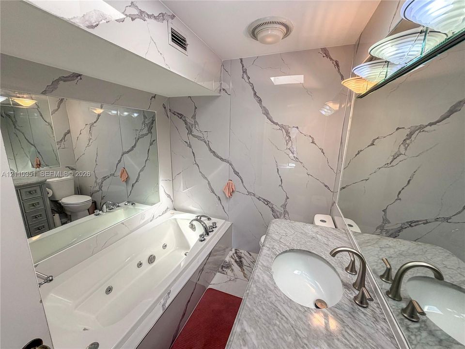 Bathroom featuring stone-finish wall cladding, a jetted soaking tub, a stone-top vanity with an integrated undermount sink, and multi-light wall-mounted fixture