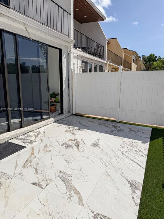 Patio featuring large-format stone-finish tiles, white vinyl privacy fencing, synthetic turf, black-framed sliding glass doors, and a second-story balcony with black metal railings
