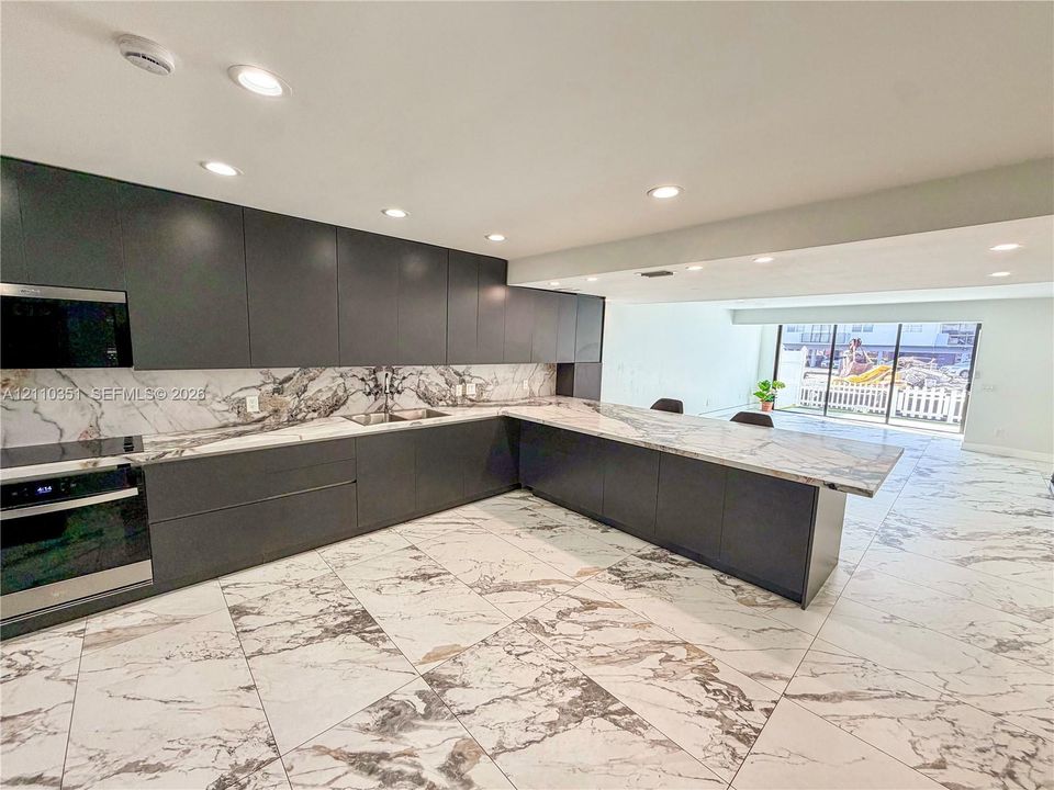 Kitchen featuring dark-toned flat-panel cabinetry, stone-patterned countertops, a matching full-height stone-patterned backsplash, recessed ceiling lighting, and large-format stone-patterned flooring