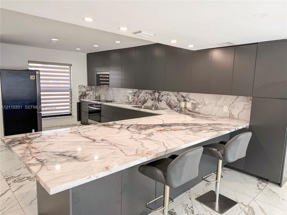 Kitchen featuring flat-panel charcoal cabinetry, patterned stone countertops, matching full-height backsplash, recessed ceiling lighting, and large-format tile flooring
