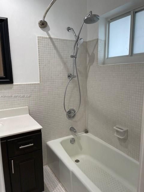Bathroom featuring a white bathtub with a dual-head chrome shower fixture, small-format neutral wall tiling, a dark wood-finish vanity with a white countertop, and a recessed window