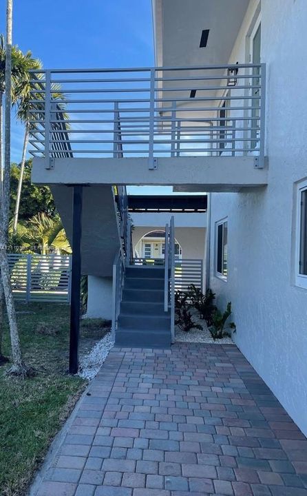 Exterior staircase with horizontal metal railing, raised balcony, paver stone walkway, and stucco wall finish