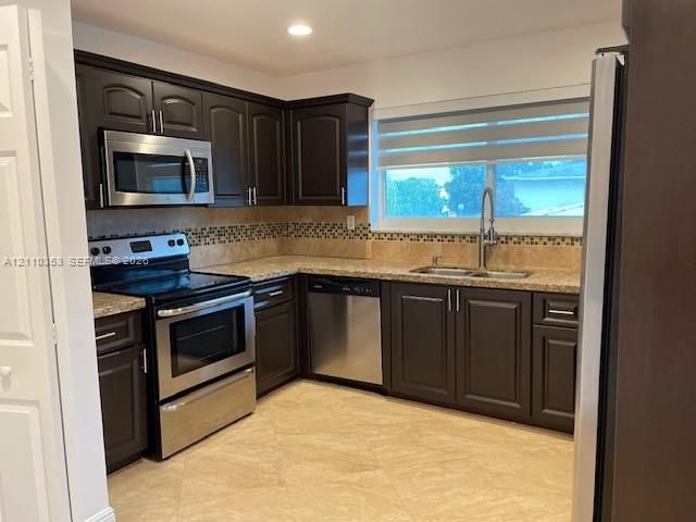 Kitchen featuring dark wood-finish cabinetry, stainless steel appliances, neutral stone-finish countertops, mosaic tile backsplash, and light-tone tile flooring