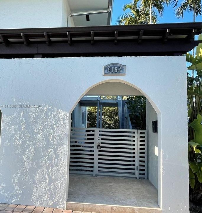 Stucco-finished entryway featuring an arched passage, horizontal slatted gate, stone tile flooring, and dark wood-finish eaves