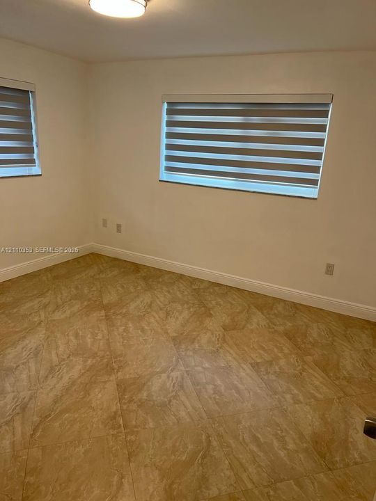 Neutral-toned interior room featuring large-format ceramic tile flooring, dual windows with banded roller shades, and a flush-mount ceiling light fixture