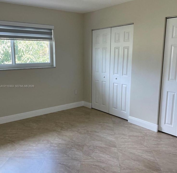 Neutral-toned room featuring large-format tile flooring, dual bi-fold closet doors, and a single window with a roller shade