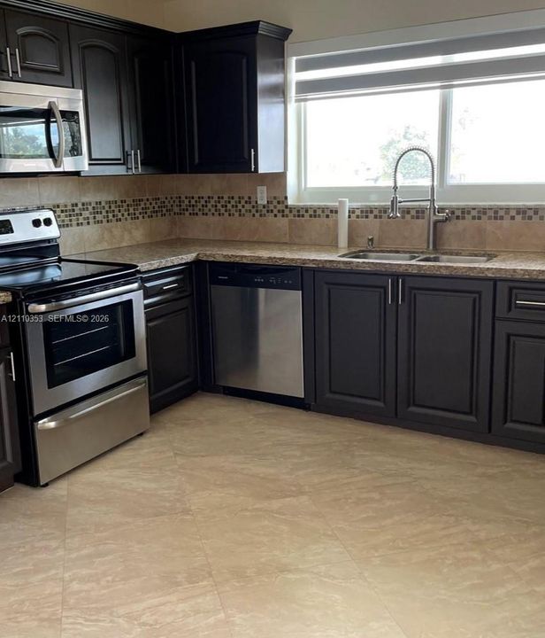 Kitchen featuring dark wood cabinetry, stainless steel appliances, stone-look countertops, a mosaic tile backsplash, and ceramic floor tiling