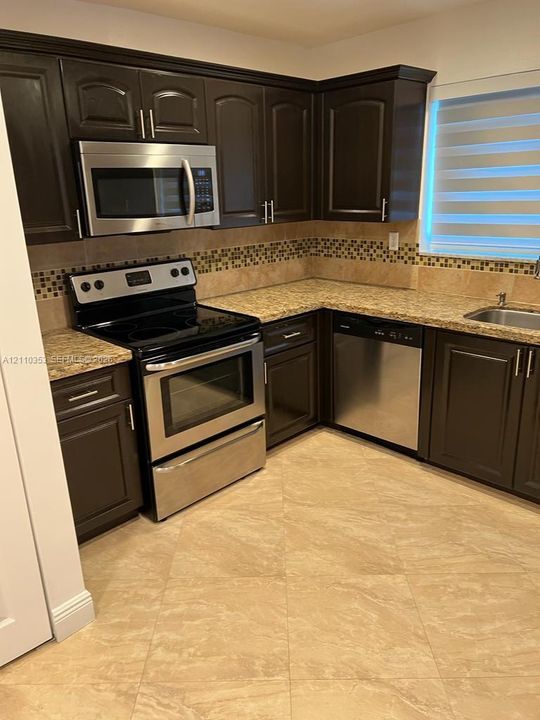 Kitchen featuring dark-finish cabinetry, stainless steel appliances, stone-look countertops, decorative tile backsplash, and ceramic tile flooring
