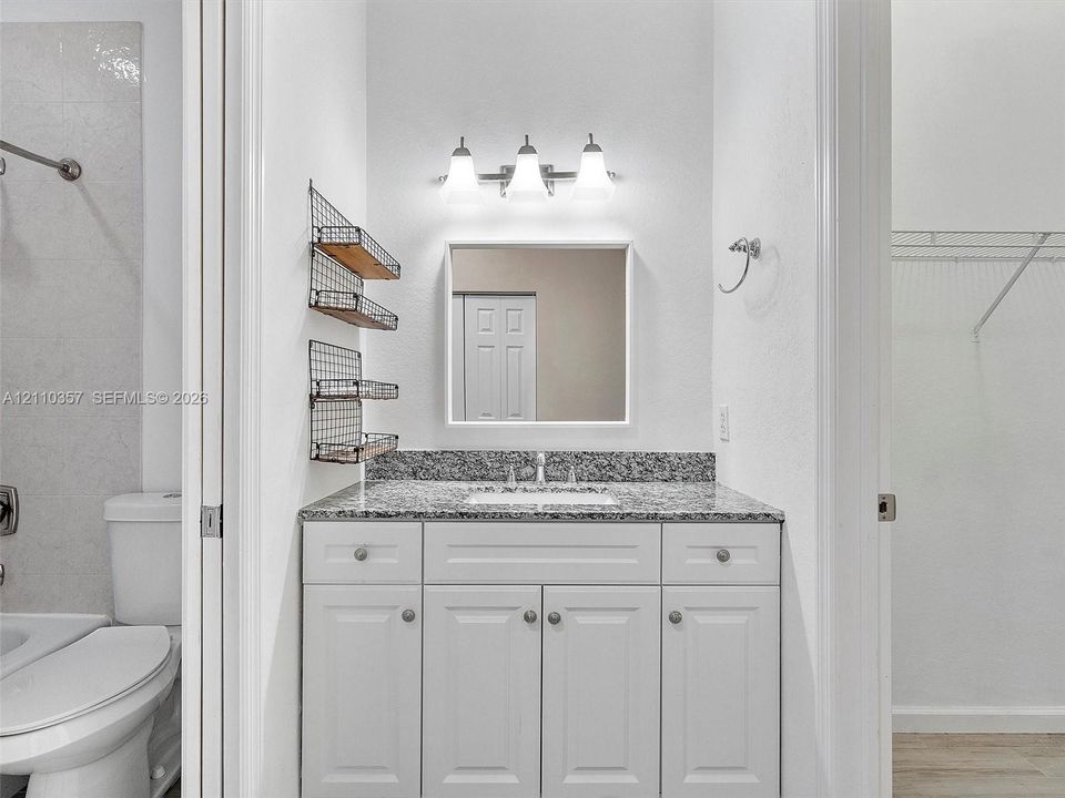 White cabinetry vanity with stone-finish countertop, undermount sink, three-light fixture, and square wall-mounted mirror