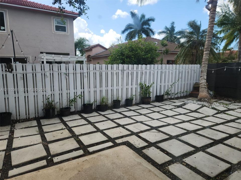 Backyard patio featuring geometric concrete pavers with dark gravel inlay, enclosed by a white privacy fence and a mature palm tree