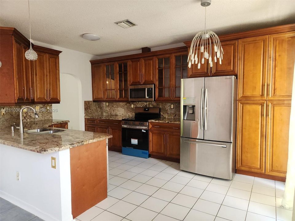 Kitchen featuring medium-tone wood cabinetry, stone countertops, stainless steel appliances, tile flooring, and decorative pendant lighting