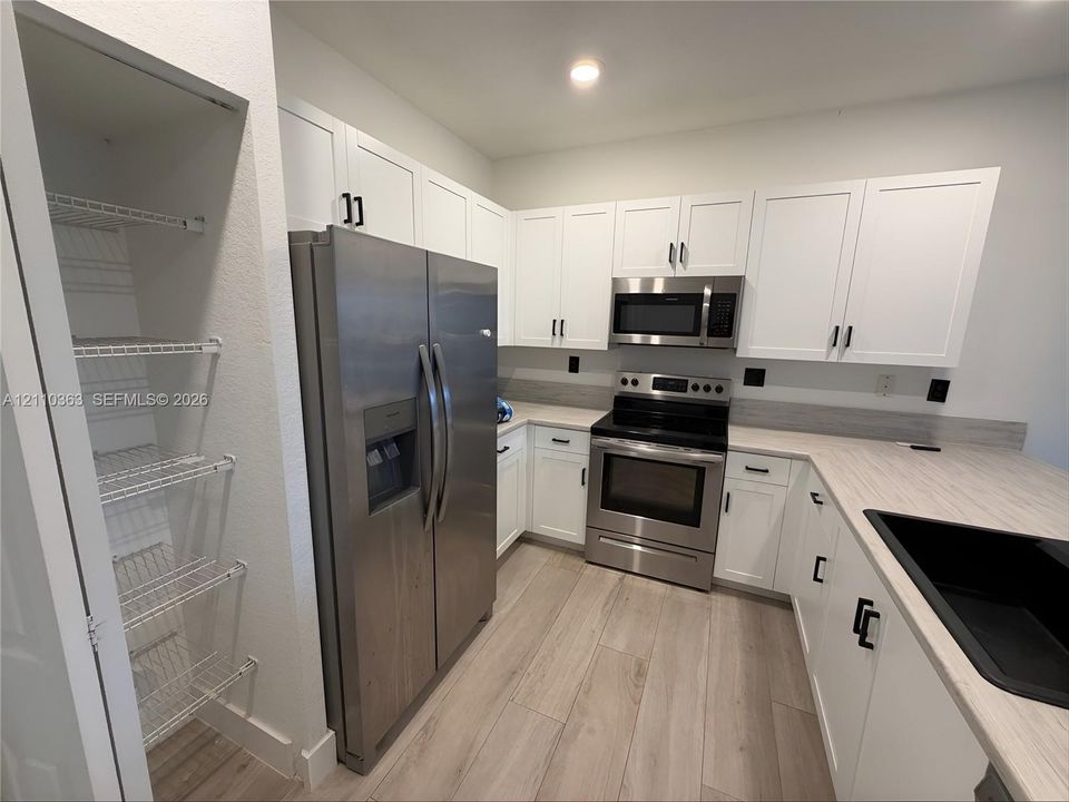 Kitchen featuring white shaker-style cabinetry, stainless steel appliances, wood-finish flooring, a built-in wire shelving pantry, and a matte black undermount sink