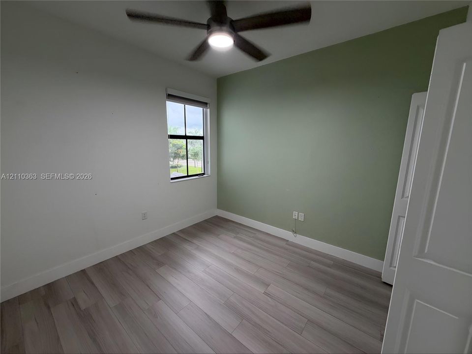 Room featuring wood-finish flooring, a sage green accent wall, a single-pane window with dark framing, and an integrated ceiling fan with light fixture