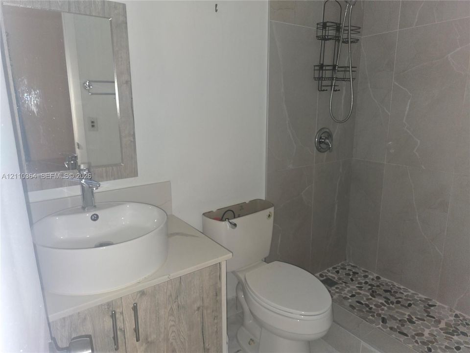 Bathroom featuring a vessel sink on a wood-finish vanity, polished faucet, framed mirror, and a shower with stone-tile surround and pebble-texture flooring