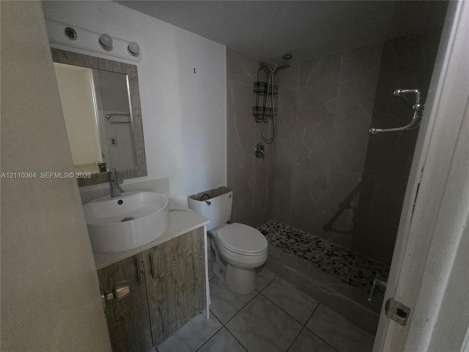 Bathroom featuring a vessel sink with a single-handle faucet, wood-finish cabinetry, a framed mirror with integrated light fixture, tiled shower surround, and pebble-textured shower floor