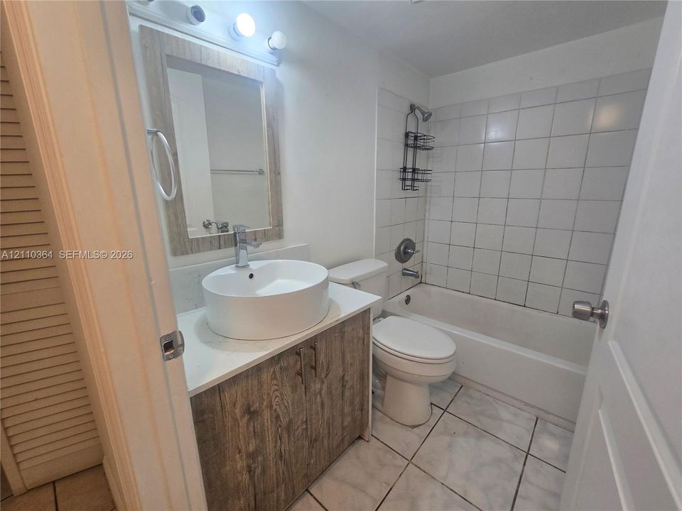 Bathroom featuring a wood-finish vanity with a rectangular vessel sink, solid-surface countertop, tile flooring, and a shower-tub combination with white wall tile