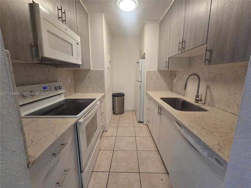Galley kitchen featuring grey wood-finish cabinetry, stone-patterned countertops, a stainless steel undermount sink with a high-arch faucet, and ceramic tile flooring