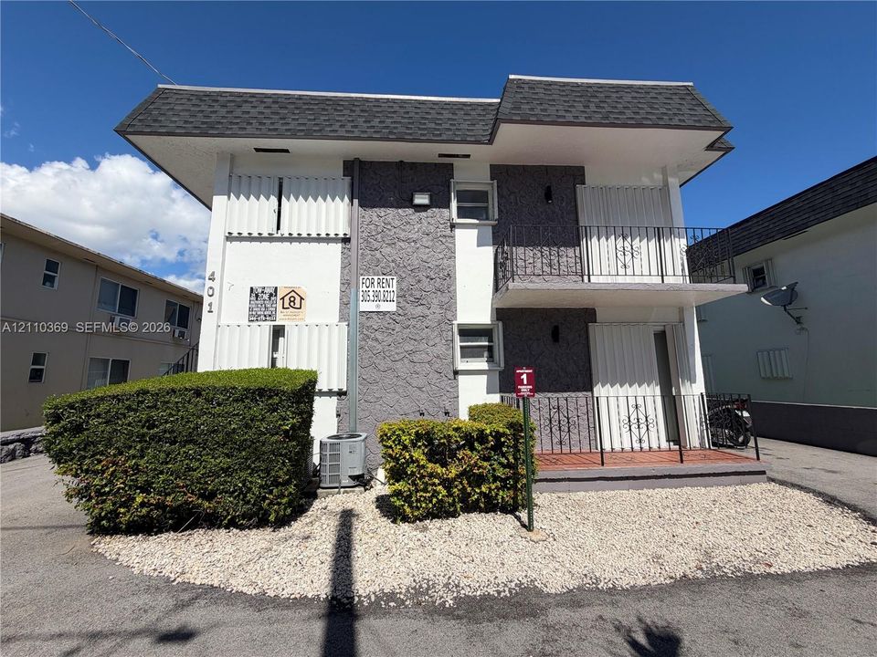 Two-story multi-family building featuring a textured stucco and smooth-finish exterior, dual private balconies with decorative metal railings, shingle roofing, and landscaped ground-level shrubbery
