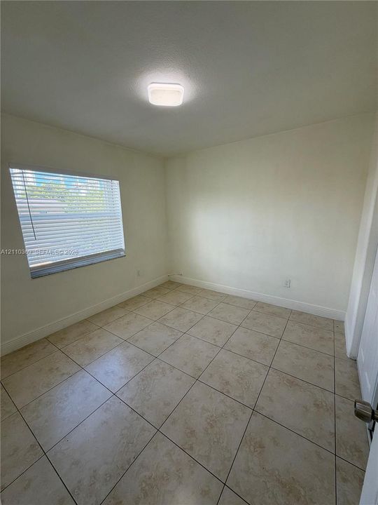 Neutral-toned interior space featuring light-colored square floor tiles, a single-pane window with horizontal blinds, and a central flush-mount ceiling light fixture