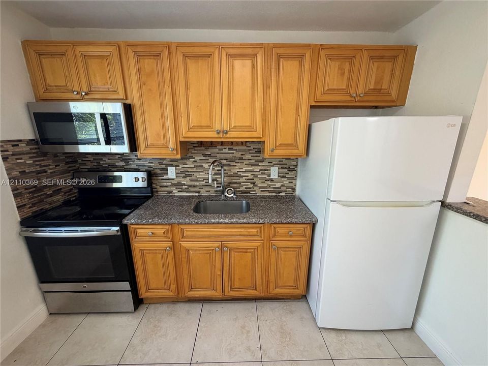 Kitchen featuring natural-finish wood cabinetry, a decorative multi-toned tile backsplash, stone-composite countertops, stainless steel appliances, and ceramic tile flooring