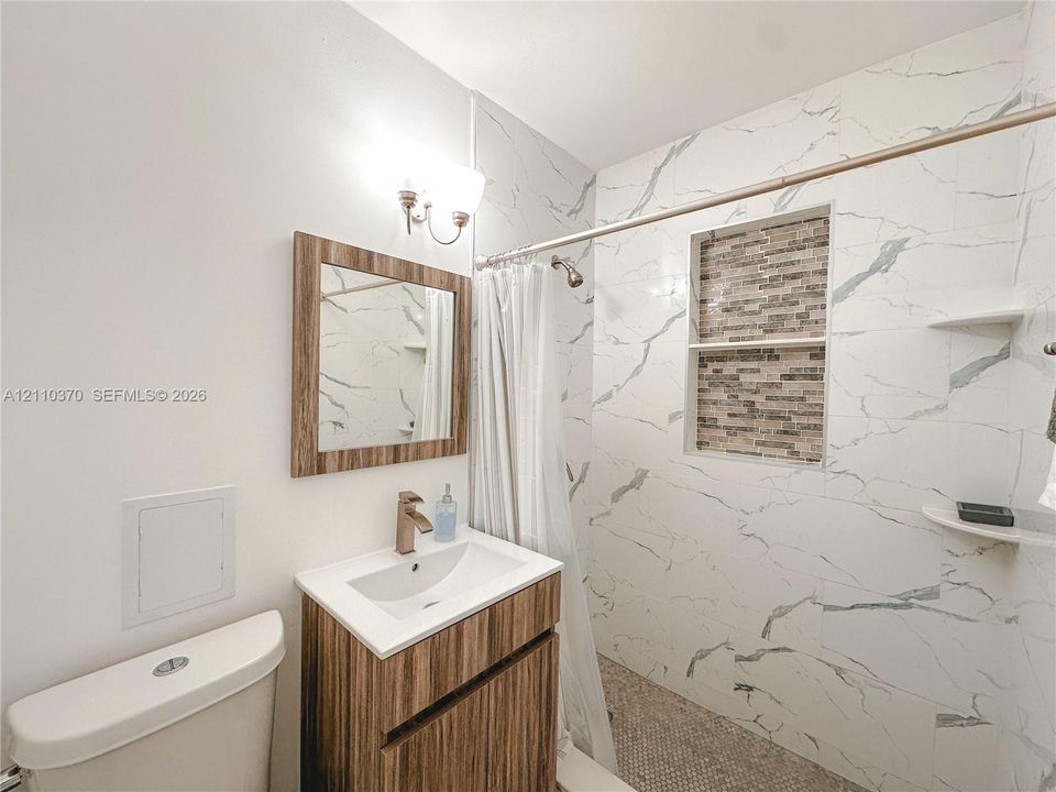 Bathroom featuring a vanity with a white countertop, wall-mounted light fixture, mirror with a wood-finish frame, and a shower area with large-format tile surround, stone-finish tile accent niche, and built-in shelving