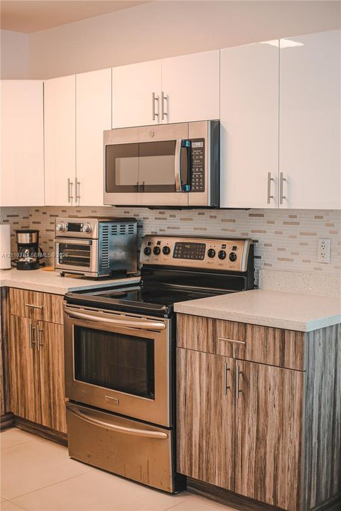 Kitchen featuring high-gloss white upper cabinetry, wood-finish lower cabinetry, stone-composite countertops, stainless steel appliances, and a linear tile backsplash