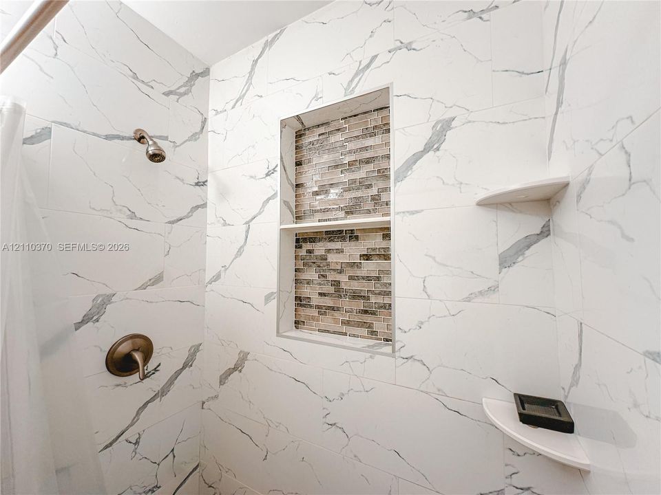 Shower surround finished with large-format white veined tile, a multi-tier built-in tiled niche with decorative mosaic detailing, two integrated corner shelves, and a metal finish wall-mounted shower fixture