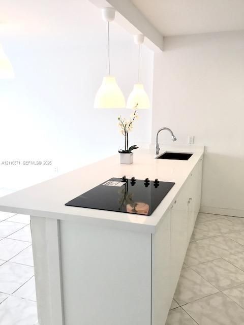 Kitchen island featuring white countertops, an integrated glass cooktop, an undermount sink with a gooseneck faucet, dual pendant lighting, and neutral tile flooring