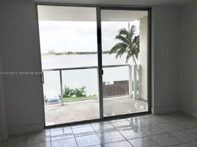 Floor-to-ceiling sliding glass door, tiled flooring, and balcony with metal and glass railing overlooking a waterfront and palm tree backdrop