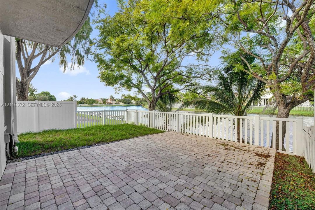 Paver patio area featuring a white vinyl fence, green lawn, mature trees, and water frontage
