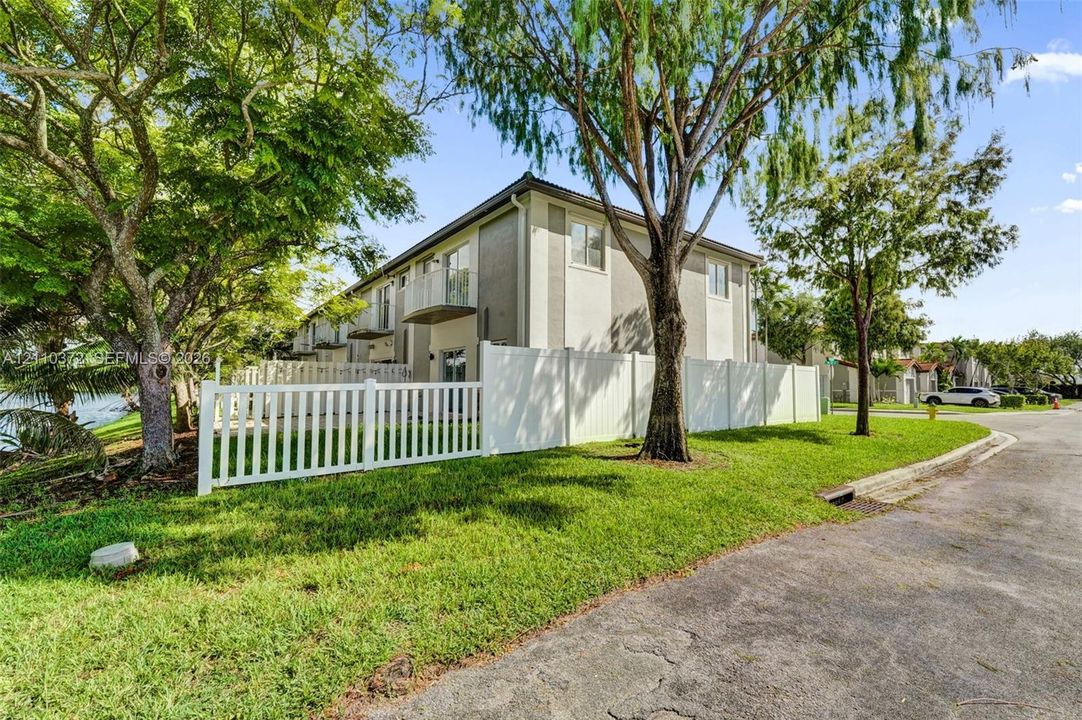 Multi-story building exterior featuring smooth stucco siding, white vinyl perimeter fencing, private balconies, and mature landscaping
