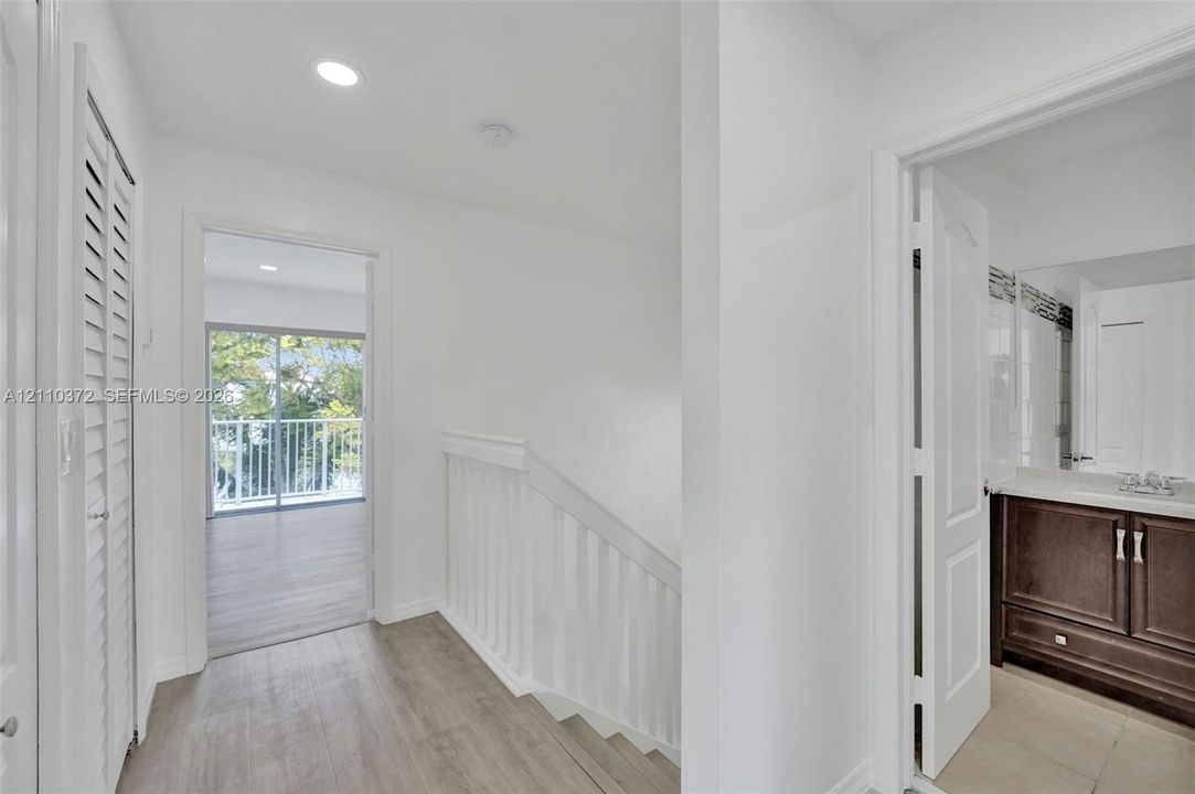 Interior hallway featuring wood-finish flooring, white louvered closet doors, recessed lighting, a white wooden stair railing, and a bathroom vanity with dark wood-finish cabinetry