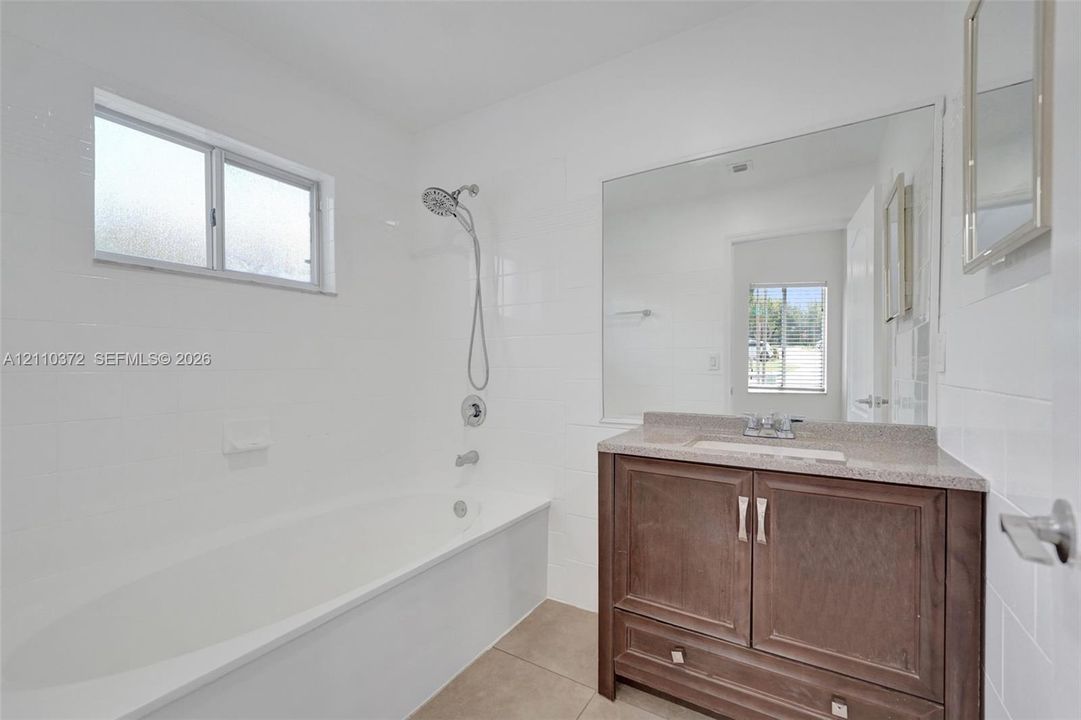 Bathroom featuring a deep soaking tub with a handheld shower fixture, full-width wall mirror, wood-finish vanity with a stone-composite countertop, and white wall tile