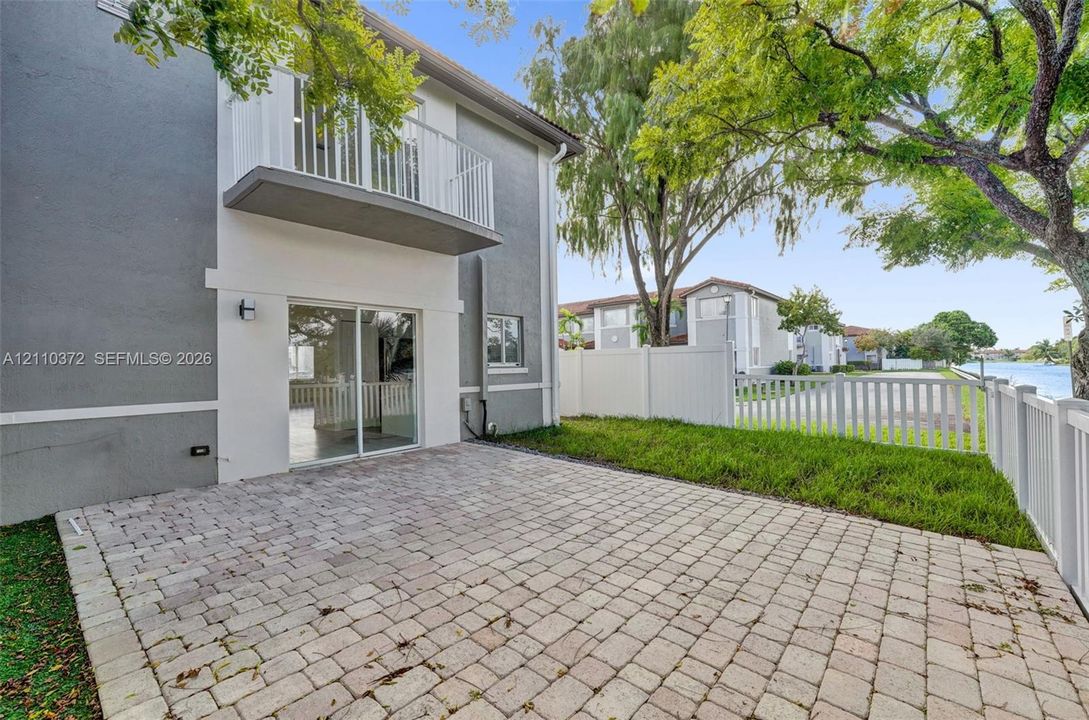 Exterior featuring a paver patio, second-level balcony with metal railing, sliding glass doors, and white vinyl privacy fencing