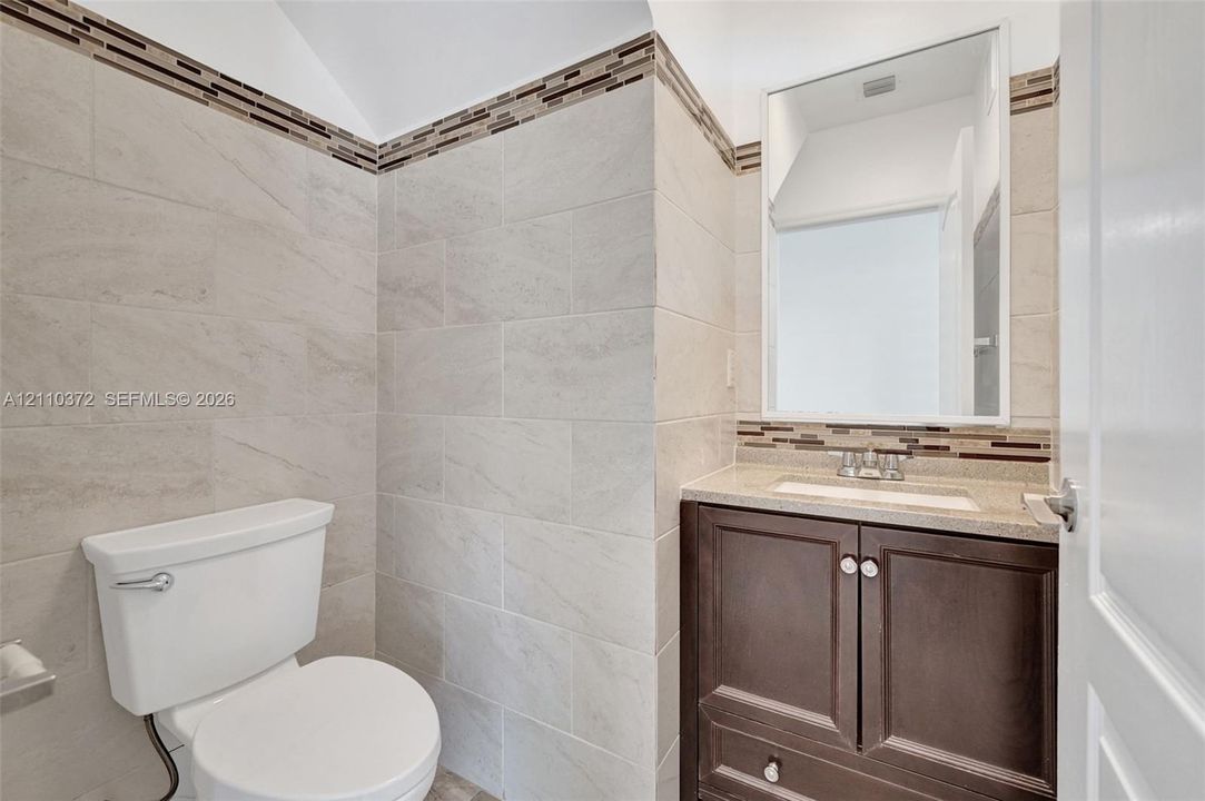 Dark wood-finish vanity with a stone-composite countertop, rectangular undermount sink, wall-mounted mirror, and floor-to-ceiling tilework featuring decorative mosaic accents