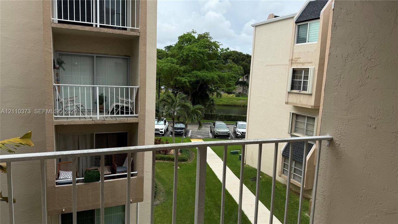 Private balcony with white metal railings, overlooking a landscaped courtyard with paved walkways, mature trees, and water frontage