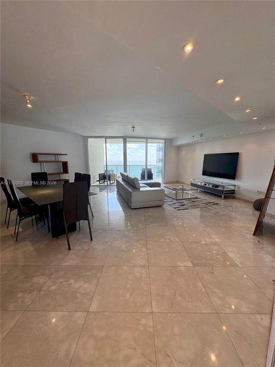 Open-concept living area featuring large-format tile flooring, recessed ceiling lighting, and floor-to-ceiling glass sliding doors leading to a balcony
