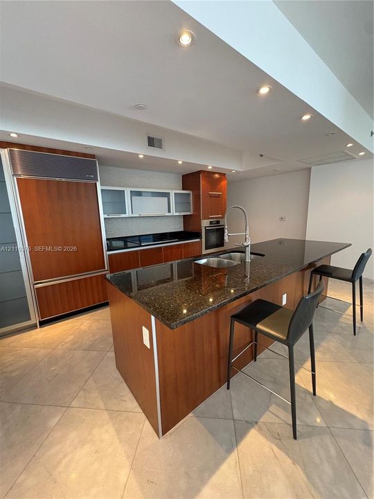 Kitchen featuring wood cabinetry, integrated refrigerator, dark stone countertops, central island with double-basin sink, and tile flooring