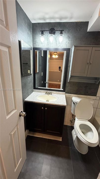 Bathroom featuring a vanity with a light-toned solid surface countertop and undermount sink, dark-finished lower cabinetry, wall-mounted upper storage, a framed mirror, and a three-light fixture